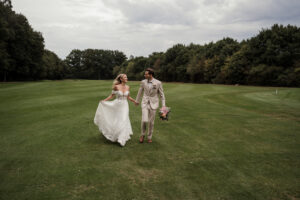 Braut und Bräutigam rennen Hand in Hand über einen Golfplatz